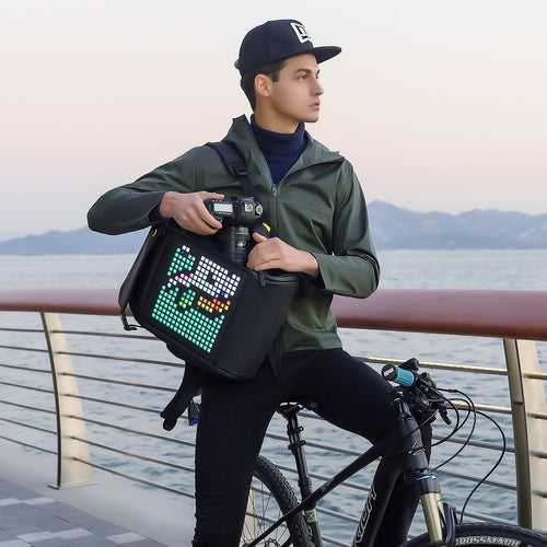 Divoom Pixoo Backpack-M mit Kamera-Pixelmotiv am Wasser, getragen von einem Mann neben seinem Fahrrad; Kamera und Fahrrad nicht enthalten